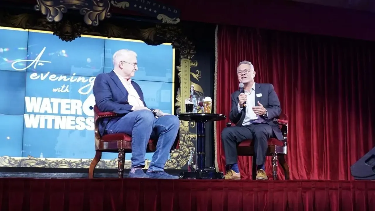 Two people seated onstage in front of an An Evening with Water Witness screen, one speaking into a microphone.