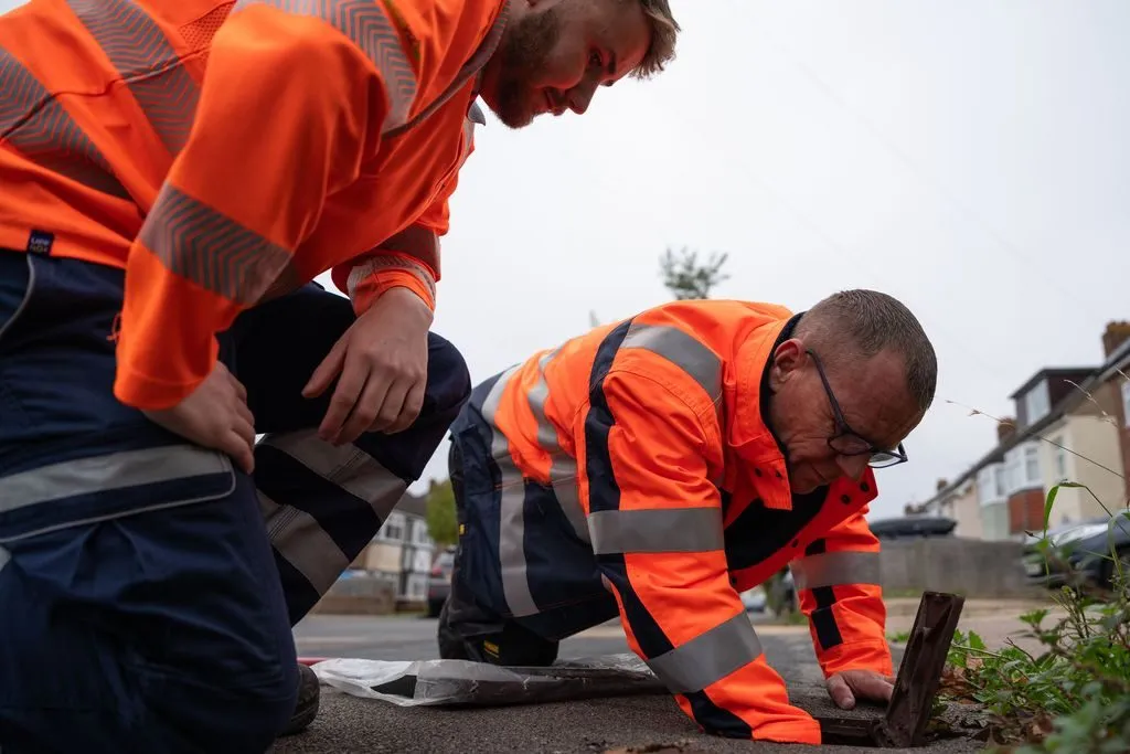 Two Portsmouth Water technicians in orange hi-vis kneel at an open pavement cover on a residential street, checking for a leak.